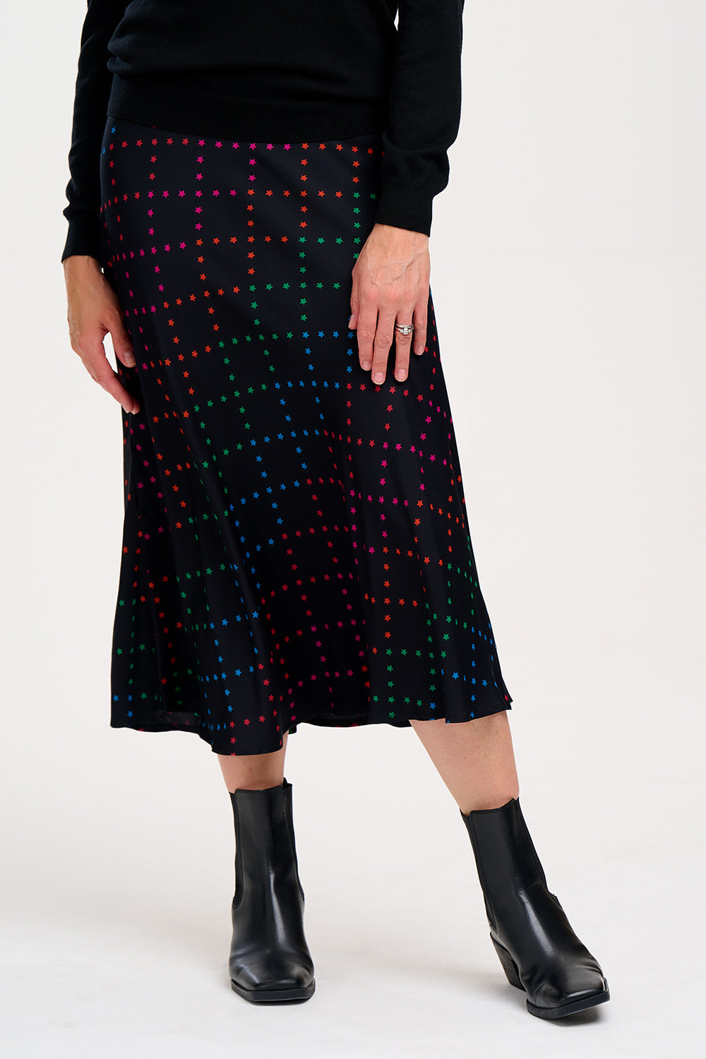 A woman wearing a midi skirt with a rainbow star lattice print in black, paired with a black top and boots. The skirt has a midi length and an elasticated waist.