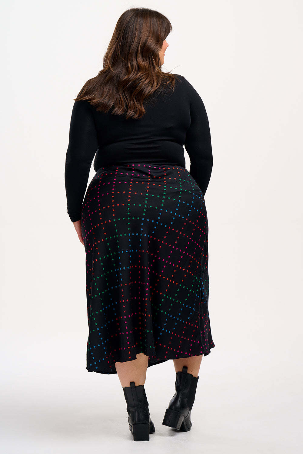 A woman wearing a midi skirt with a rainbow star lattice print in black, paired with a black top and boots. The skirt has a midi length and an elasticated waist.