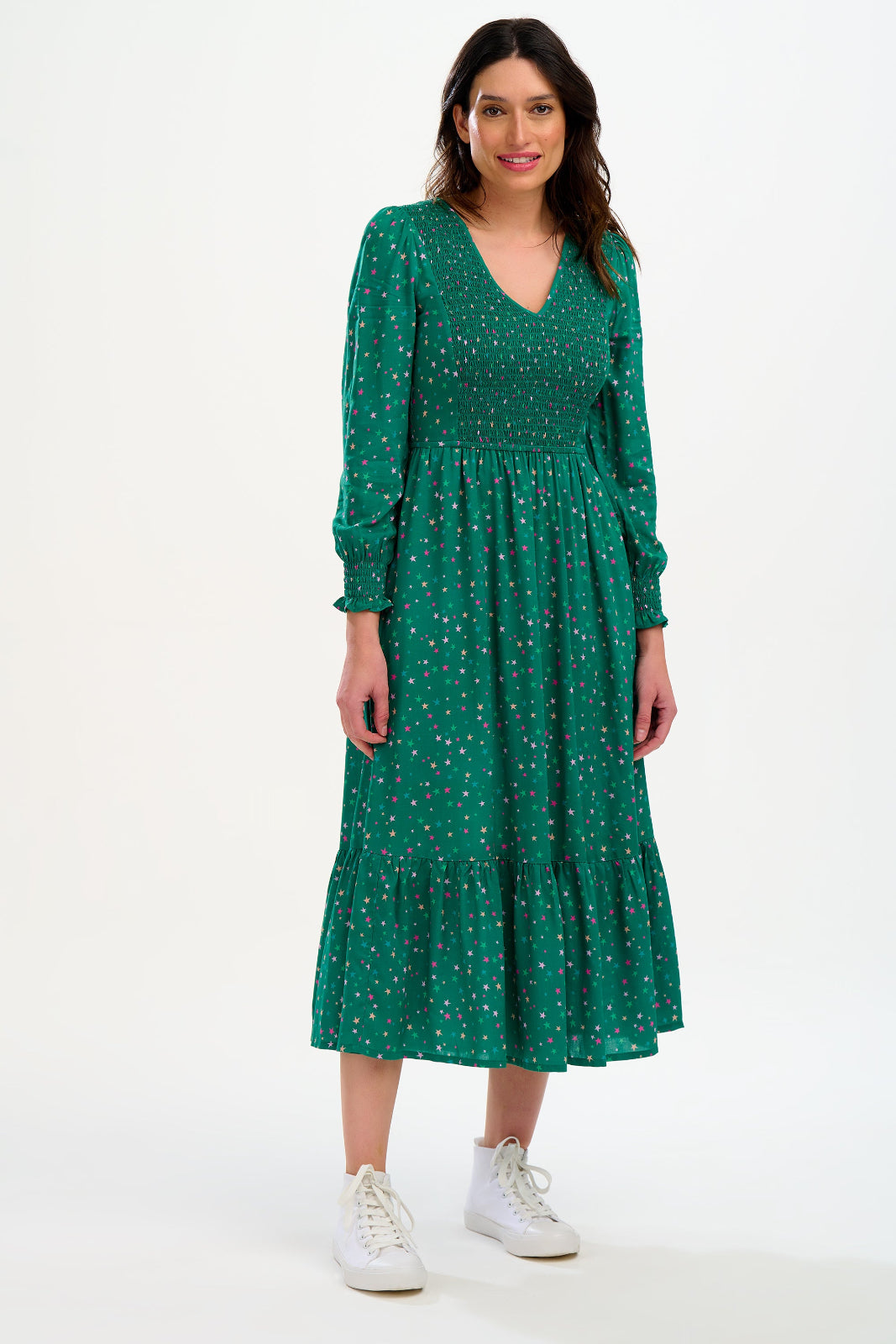 A woman standing and smiling, wearing a green mini dress with a shirred bodice and v-neckline, and long sleeves with frill cuffs. She is also wearing white sneakers.