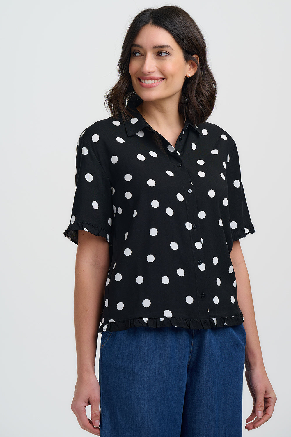 Woman wearing a black blouse with white polka dots and blue pants on a light gray background