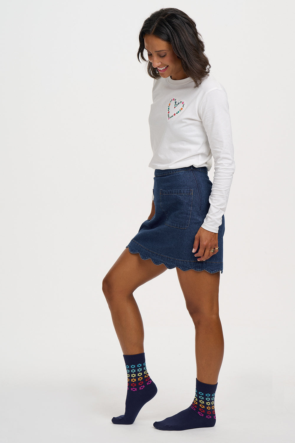 A woman modeling a white long-sleeve crewneck top with a small heart graphic, a dark blue denim mini skirt with a scalloped hem, and navy blue socks with a colorful geometric pattern, set against a plain white studio background.