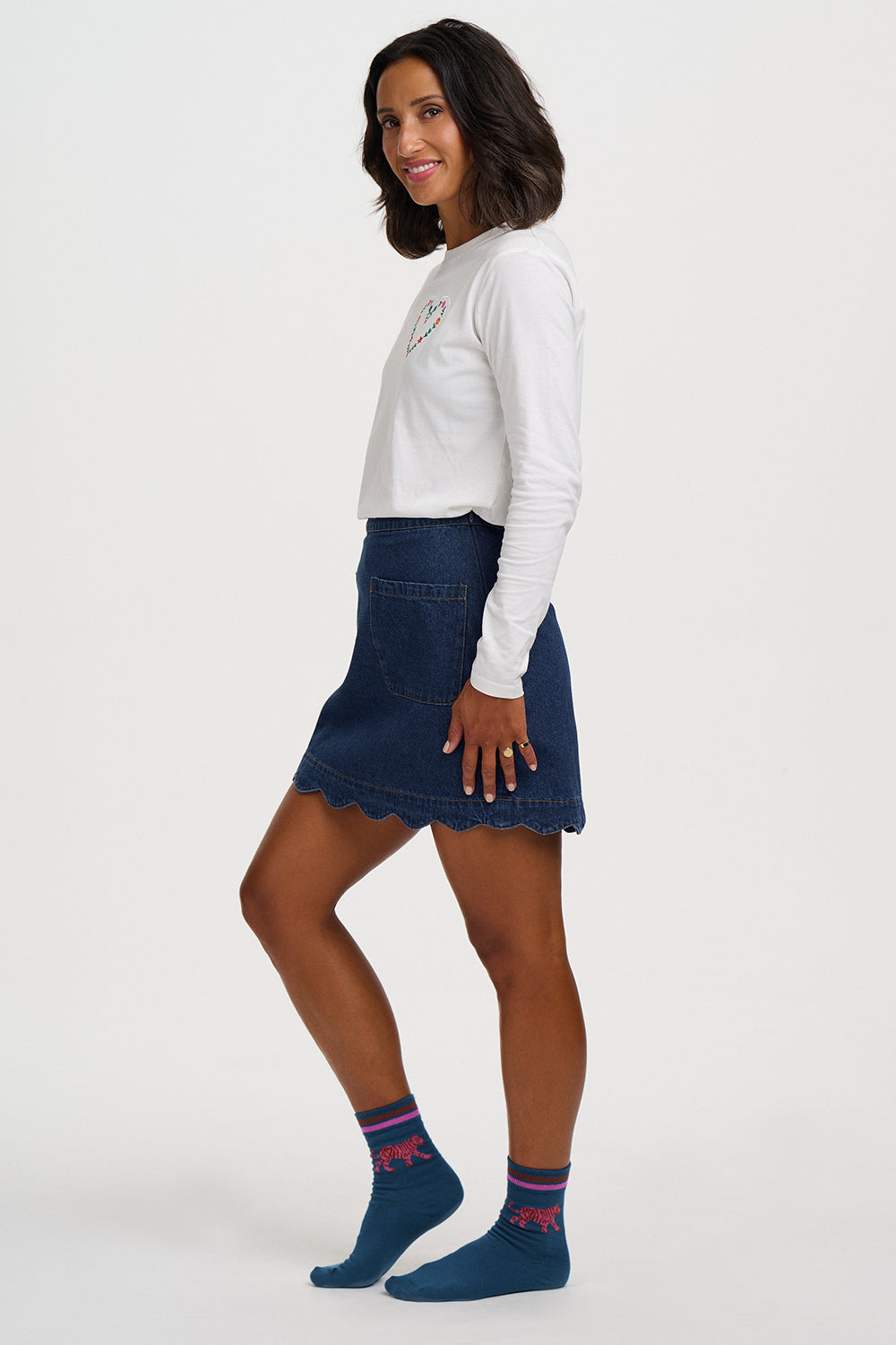 A woman modeling a dark blue denim mini skirt with a scalloped hem, paired with a white long-sleeve top and blue socks, against a plain light gray background.