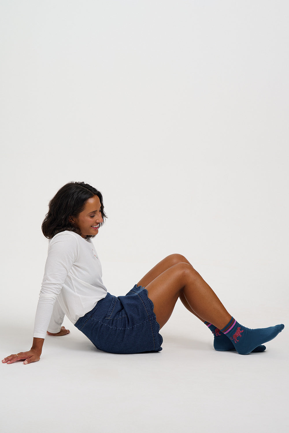 A woman sitting on a white studio floor wearing a white long-sleeve top, dark blue shorts, and dark blue crew socks with a red logo.