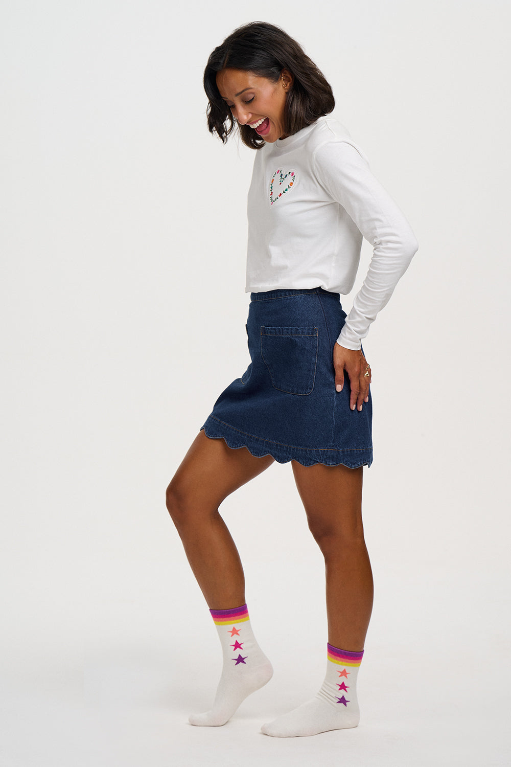 A woman modeling a white long-sleeve turtleneck top, a blue denim mini skirt with a scalloped hem, and white socks with colorful stripes and stars, posed against a solid light gray background.