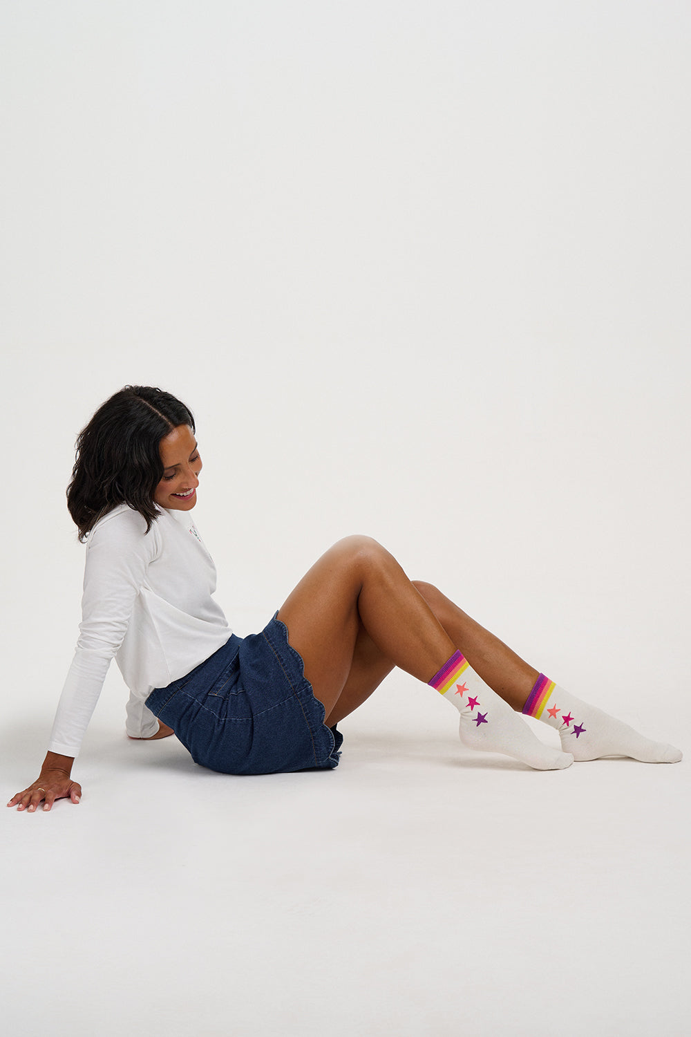 A woman sitting on a white studio floor wearing white crew socks with colorful star patterns, a white long-sleeve top, and navy blue shorts.