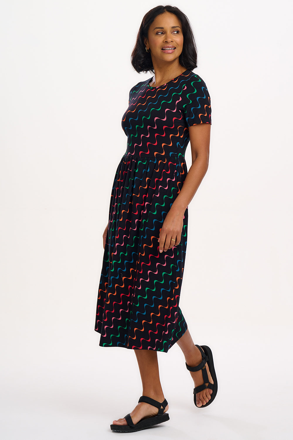 A model wearing a midi-length jersey cotton dress with a round neckline, elasticated waistband, short sleeves, and a pattern of undulating waves in black, red, and green.