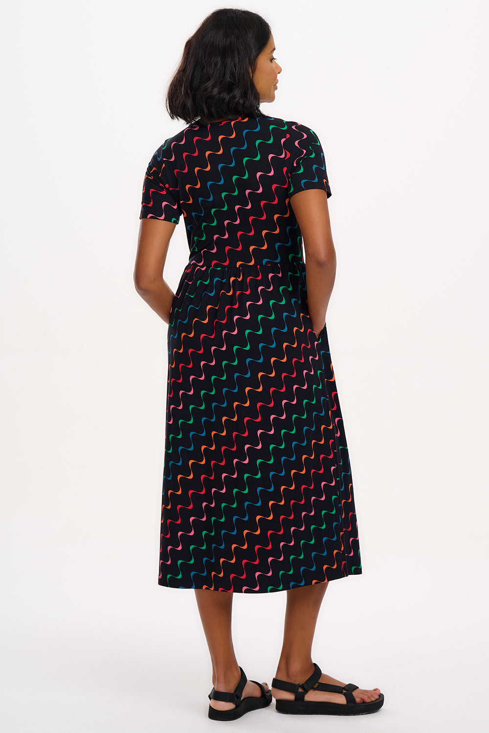 A model wearing a midi-length jersey cotton dress with a round neckline, elasticated waistband, short sleeves, and a pattern of undulating waves in black, red, and green.