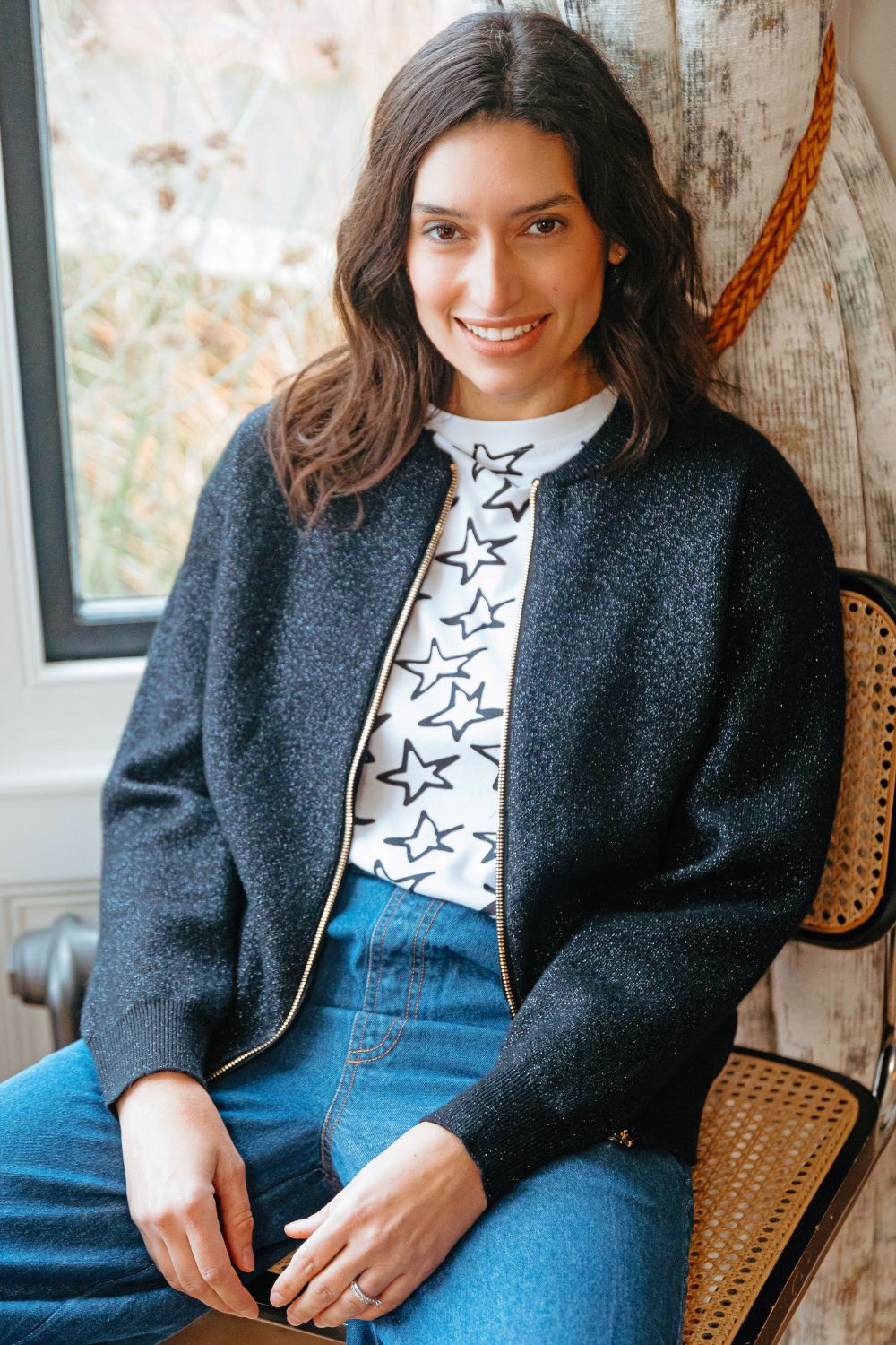 Woman sitting on a chair wearing a dark gray cardigan over a star-patterned shirt and blue jeans.