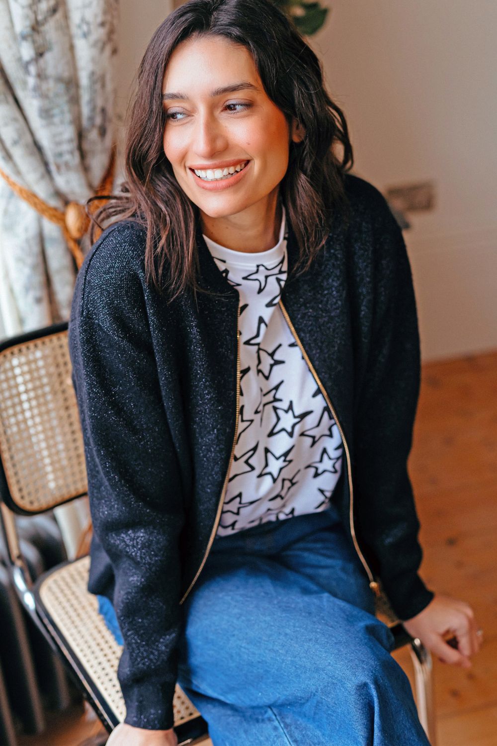 Woman wearing a black cardigan over a star-patterned shirt and blue jeans, sitting indoors.