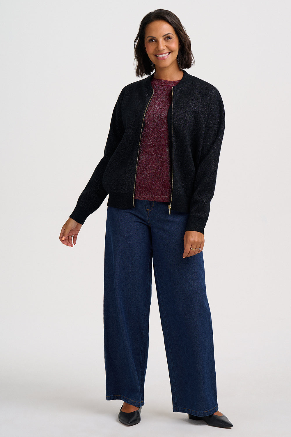 A woman modeling a black zip-up knit bomber jacket over a maroon knit top and dark blue wide-leg jeans, standing against a light gray studio background.