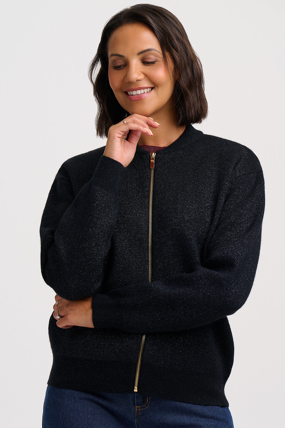 A woman wearing a dark charcoal heathered zip-up sweater with a gold zipper, paired with blue jeans, against a light gray studio background.