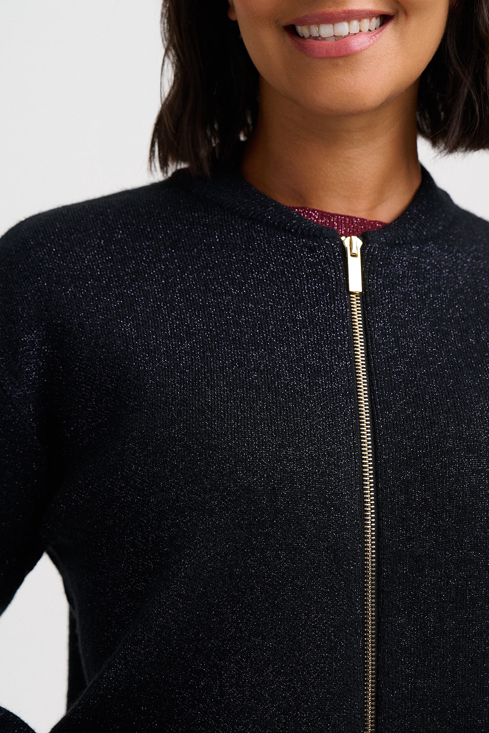 Close-up of a person wearing a dark navy or black textured zip-up sweater with a gold zipper, over a burgundy top, against a light gray background.