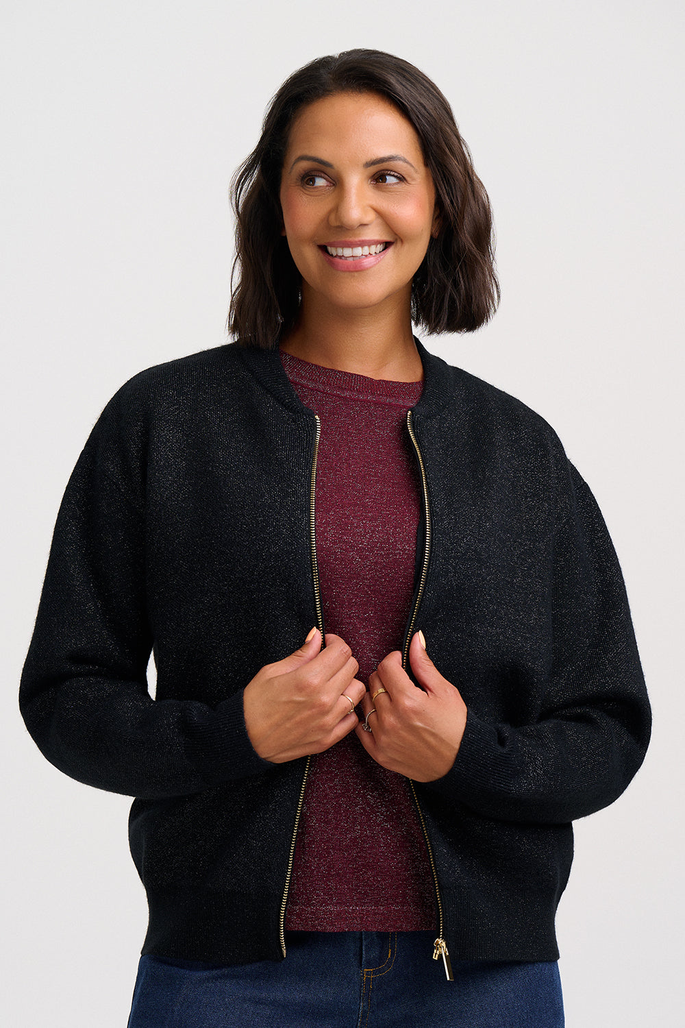 A woman wearing a dark charcoal zip-up knit jacket over a burgundy top and blue jeans, standing against a light gray studio background.