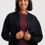 A woman wearing a dark charcoal zip-up knit jacket over a burgundy top and blue jeans, standing against a light gray studio background.