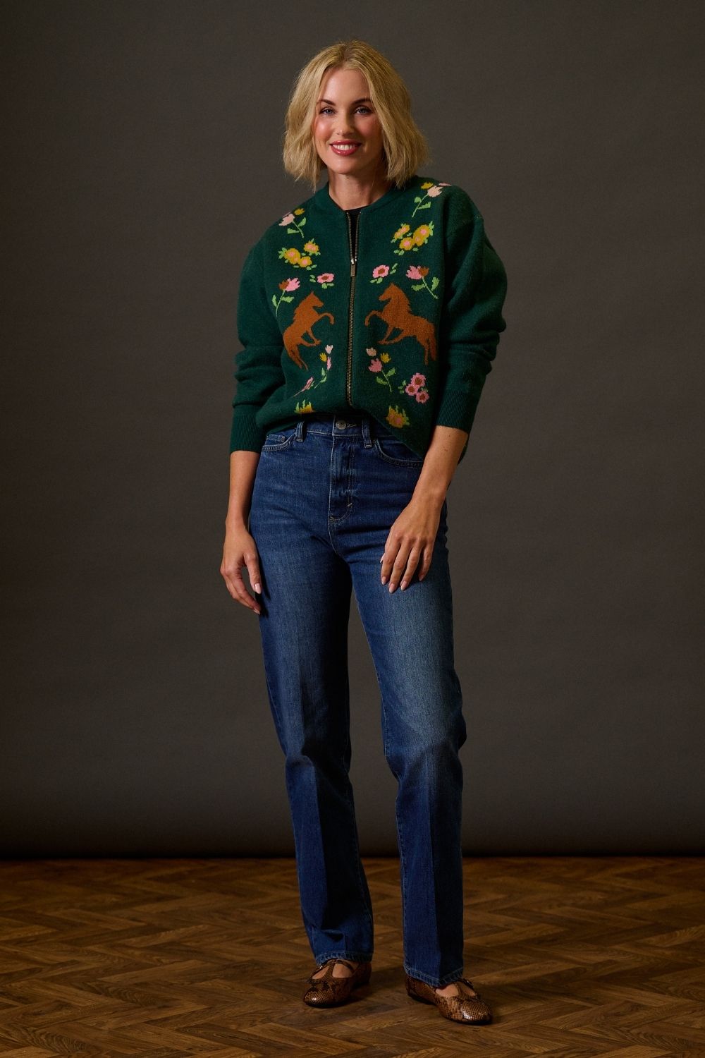 Woman wearing a green embroidered jacket and blue jeans against a dark background