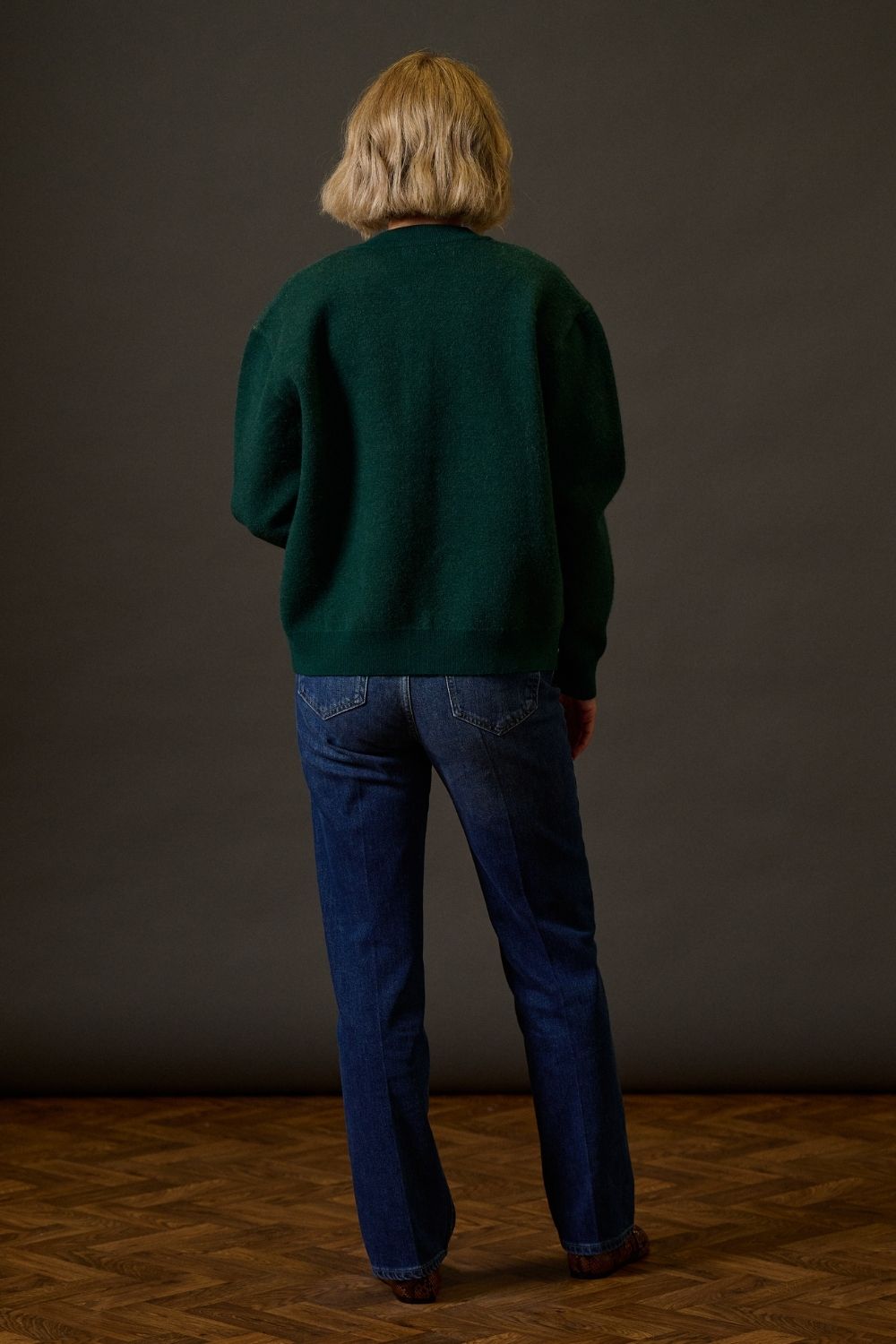 Person wearing a green sweater and blue jeans standing against a plain background