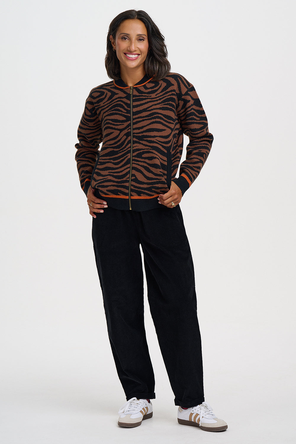 A woman modeling a brown and black tiger-striped zip-up jacket, paired with black pants and white sneakers, against a plain light gray studio background.