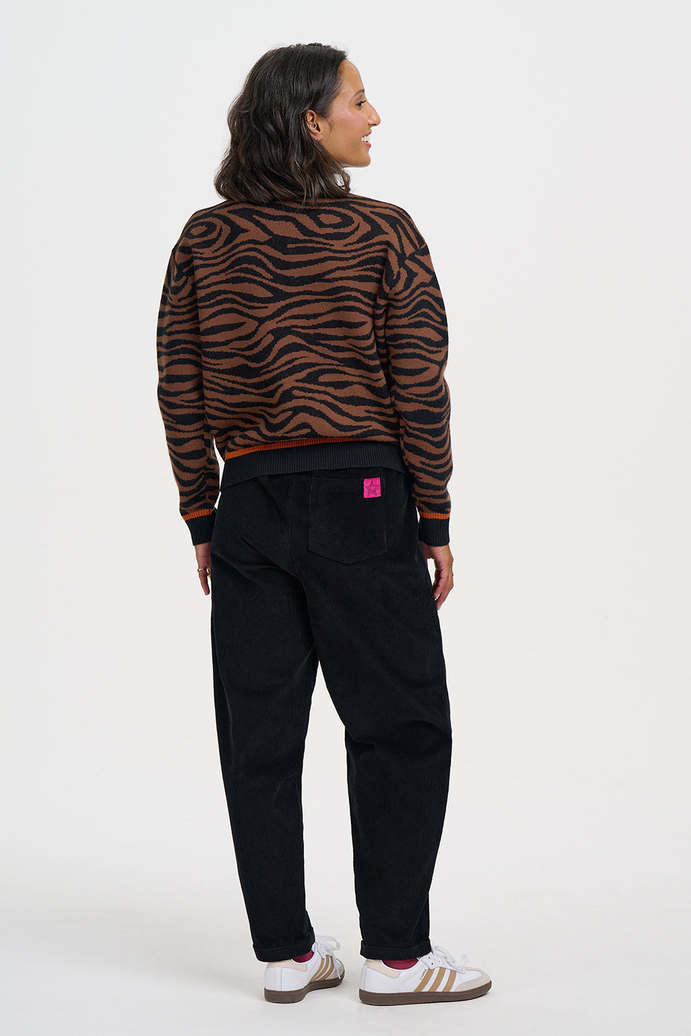 A woman shown from the back wearing a brown and black tiger-striped knit sweater, black pants, and white sneakers with gold stripes, against a plain white studio background.