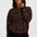 A woman wearing a brown and black tiger-stripe patterned zip-up jacket with orange ribbed trim, paired with black pants, against a light gray studio background.