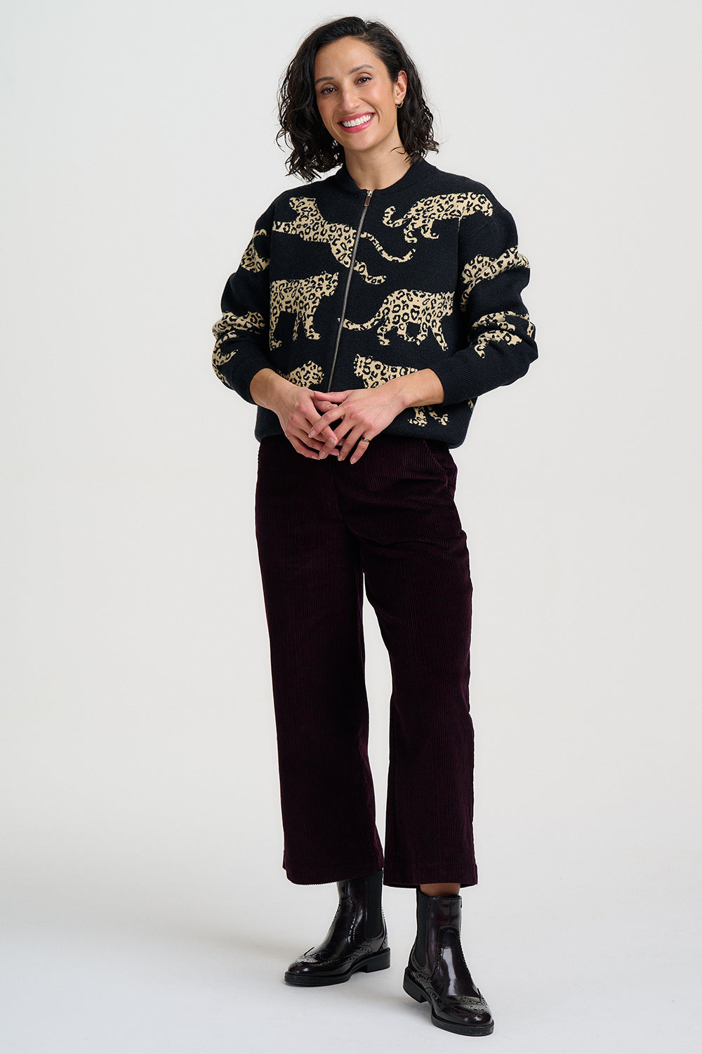 A woman modeling a black zip-up bomber jacket with gold embroidered leopard patterns, paired with dark burgundy cropped trousers and black ankle boots, against a light gray studio background.