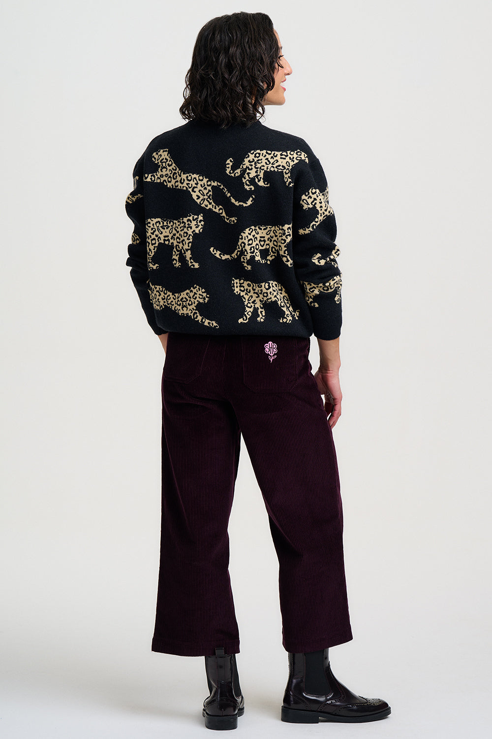 A model shown from the back wearing a black knit sweater with a gold cheetah pattern, paired with dark burgundy cropped trousers and black boots, against a light gray studio background.