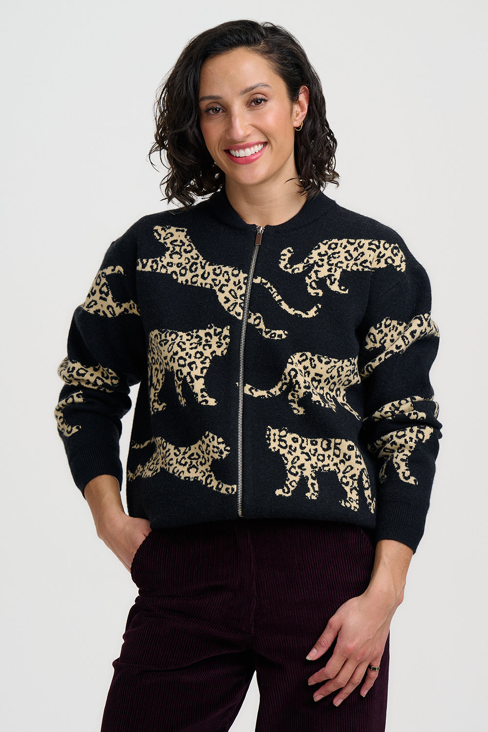 A woman wearing a black zip-up knit sweater with a beige leopard print pattern, paired with dark burgundy pants, against a light gray studio background.