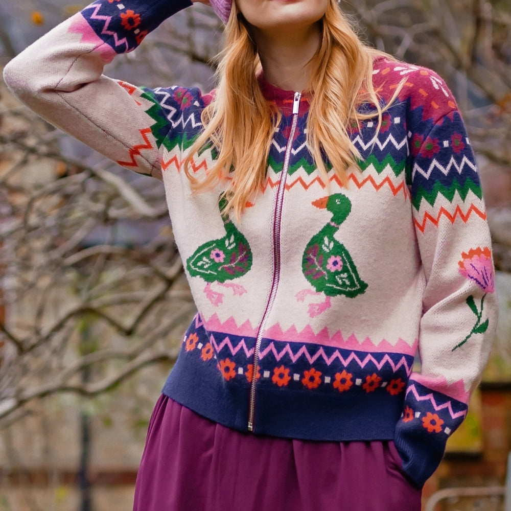 A woman wearing a colorful knit zip-up sweater with duck motifs, a pink beanie, and a purple skirt, standing outdoors in front of blurred trees.
