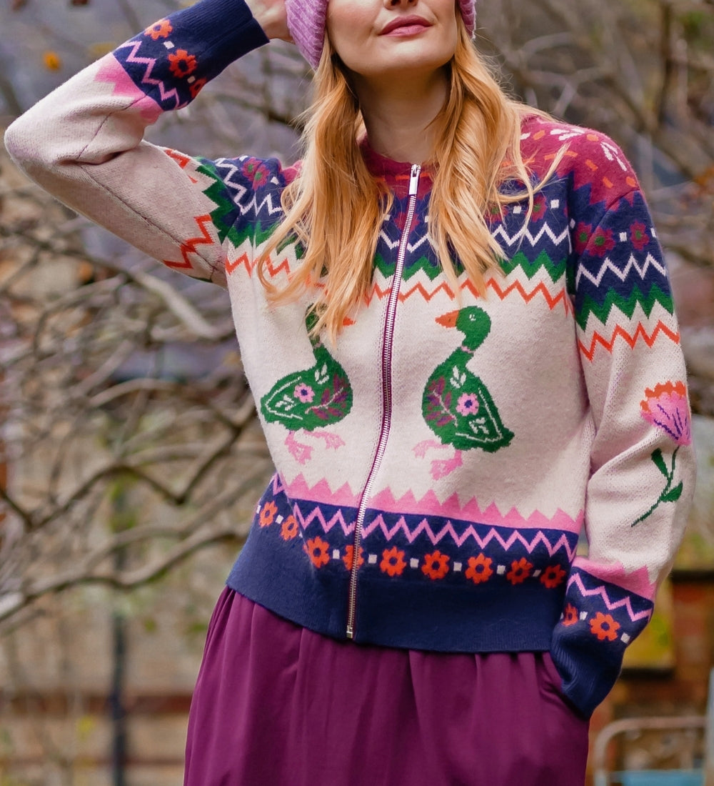 A woman wearing a colorful knit zip-up sweater with duck motifs, a pink beanie, and a purple skirt, standing outdoors in front of blurred trees.