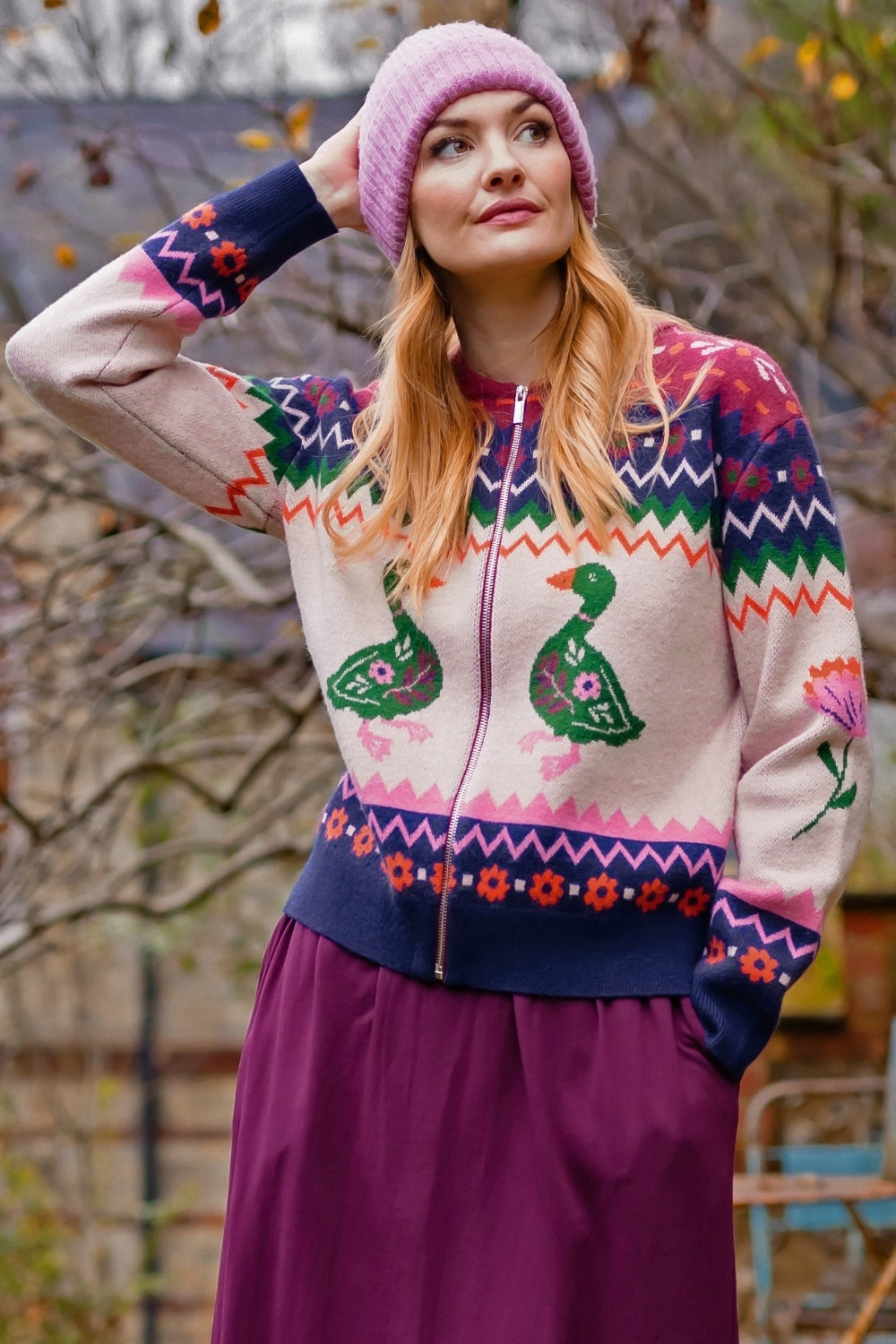 A woman wearing a colorful knit zip-up sweater with duck motifs, a pink beanie, and a purple skirt, standing outdoors in front of blurred trees.