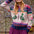 A woman wearing a colorful knit zip-up sweater with duck motifs, a pink beanie, and a purple skirt, standing outdoors in front of blurred trees.