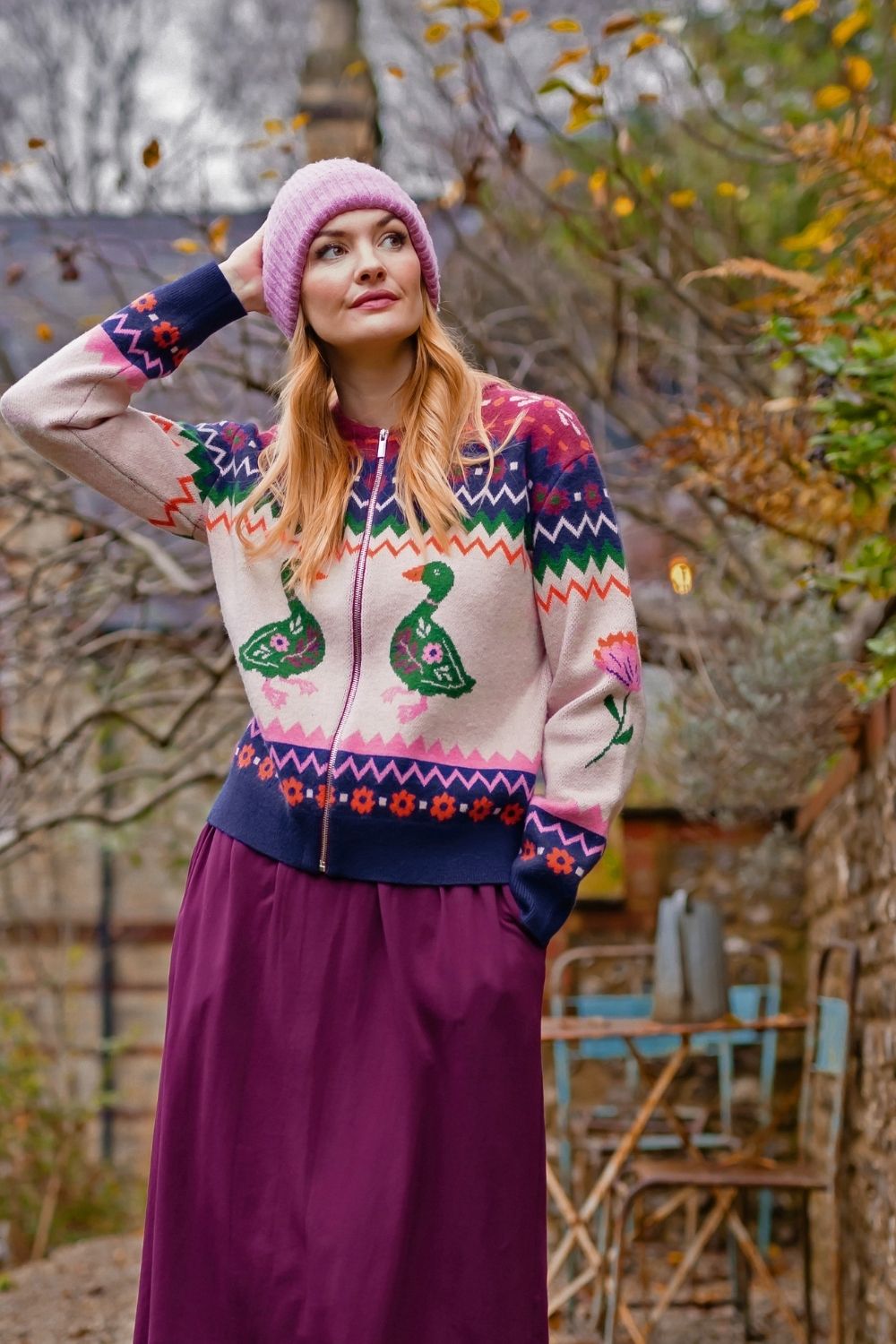 A woman wearing a colorful knit zip-up cardigan with a pink beanie and a long purple skirt, standing outdoors in a garden setting.