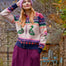 A woman wearing a colorful knit zip-up cardigan with a pink beanie and a long purple skirt, standing outdoors in a garden setting.