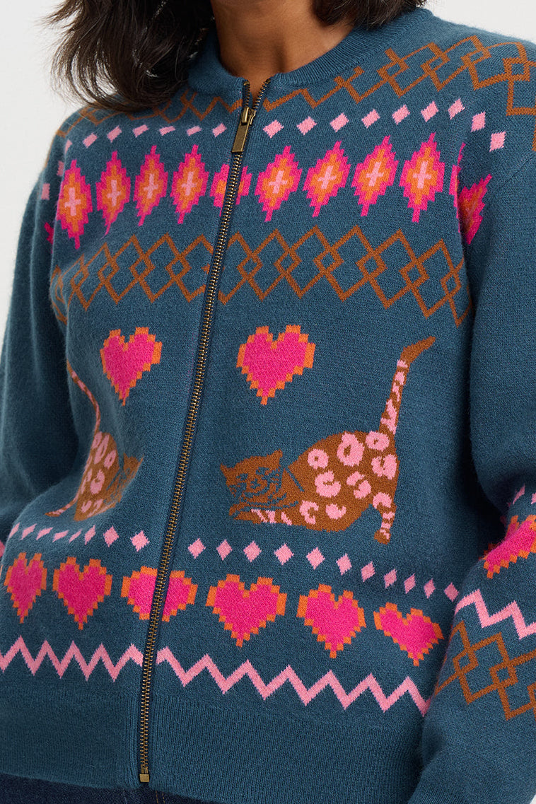 A woman wearing a teal blue knit zip-up sweater with pink and brown fair isle-style patterns, including hearts, zigzags, and cat motifs, against a light gray background.