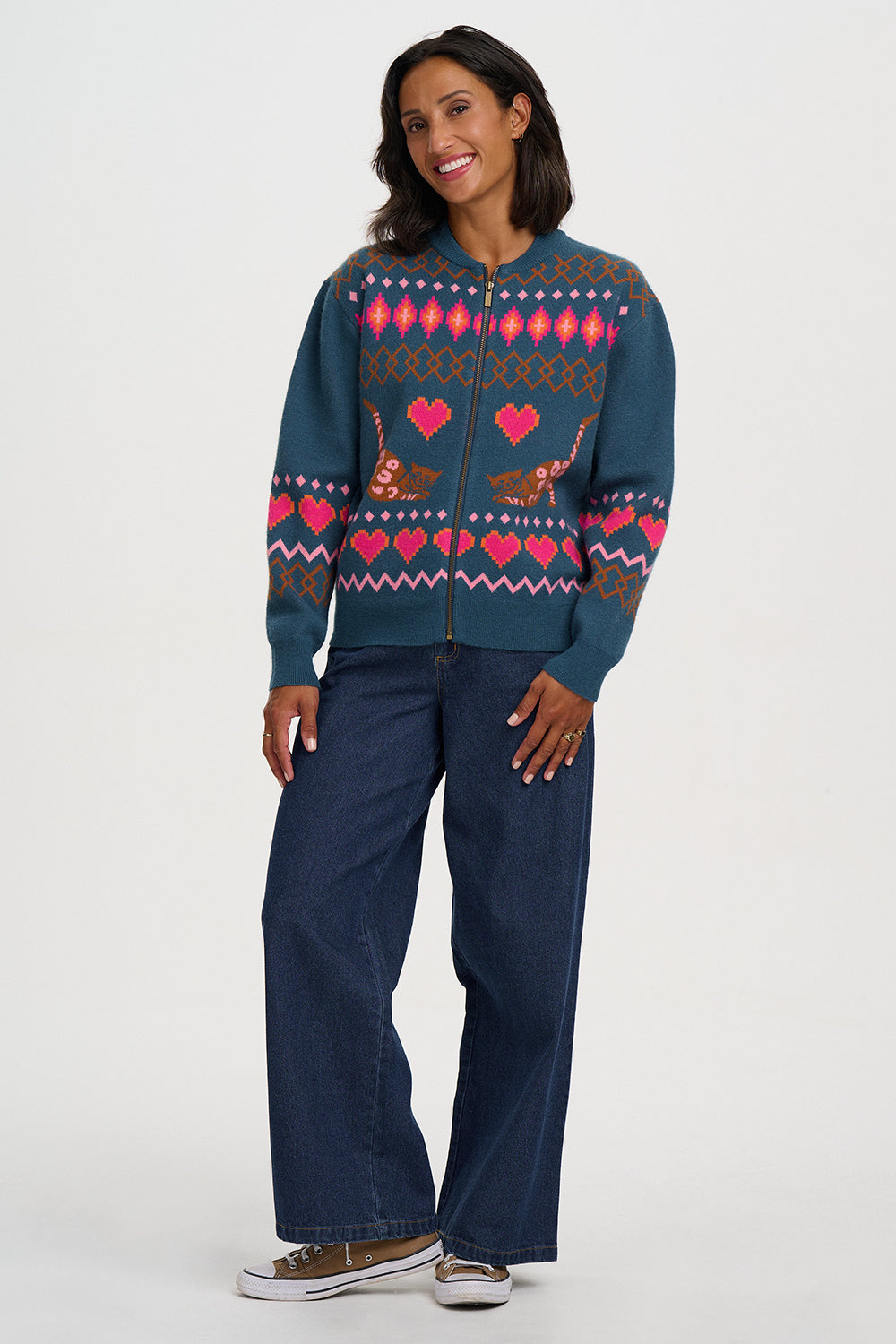 A woman modeling a teal blue zip-up sweater with a colorful Fair Isle pattern featuring pink hearts, brown reindeer, and geometric designs, paired with dark blue jeans and brown sneakers against a light gray studio background.