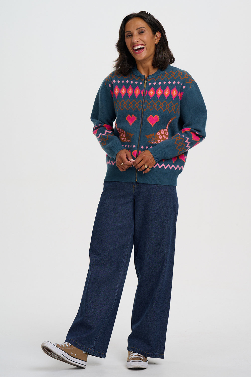 A woman modeling a dark teal zip-up sweater with a colorful Fair Isle pattern of pink hearts and birds, paired with dark blue wide-leg jeans and brown sneakers, against a light gray studio background.