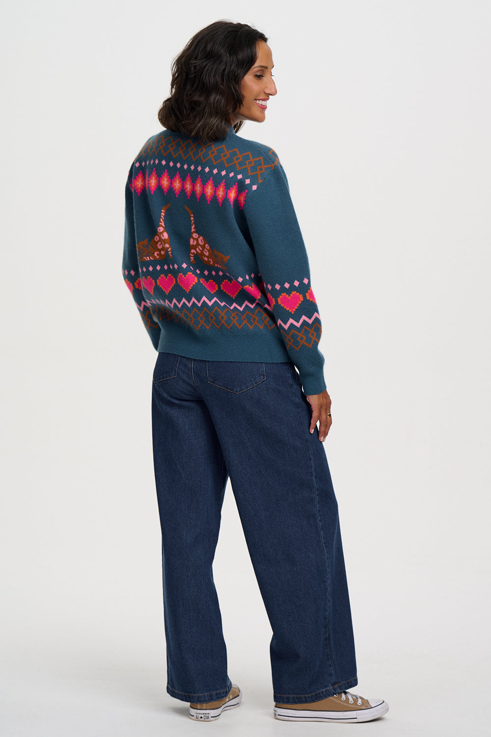 A woman shown from the back wearing a dark teal knit sweater with a colorful geometric pattern and two brown deer motifs across the upper back, paired with dark blue jeans and brown sneakers, against a light gray studio background.
