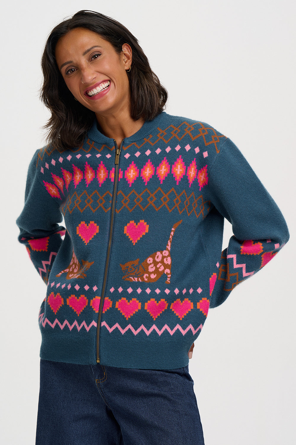 A woman wearing a teal blue zip-up sweater with a colorful knit pattern featuring pink hearts, brown cats, and geometric motifs, paired with dark blue jeans against a light gray background.
