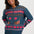 A woman wearing a teal blue zip-up sweater with a colorful knit pattern featuring pink hearts, brown cats, and geometric motifs, paired with dark blue jeans against a light gray background.
