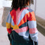 A person facing away from the camera wears a multicolored knitted bomber jacket with a geometric pattern featuring shades of navy, blue, orange, pink, and mustard.