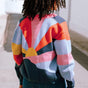 A person facing away from the camera wears a multicolored knitted bomber jacket with a geometric pattern featuring shades of navy, blue, orange, pink, and mustard.