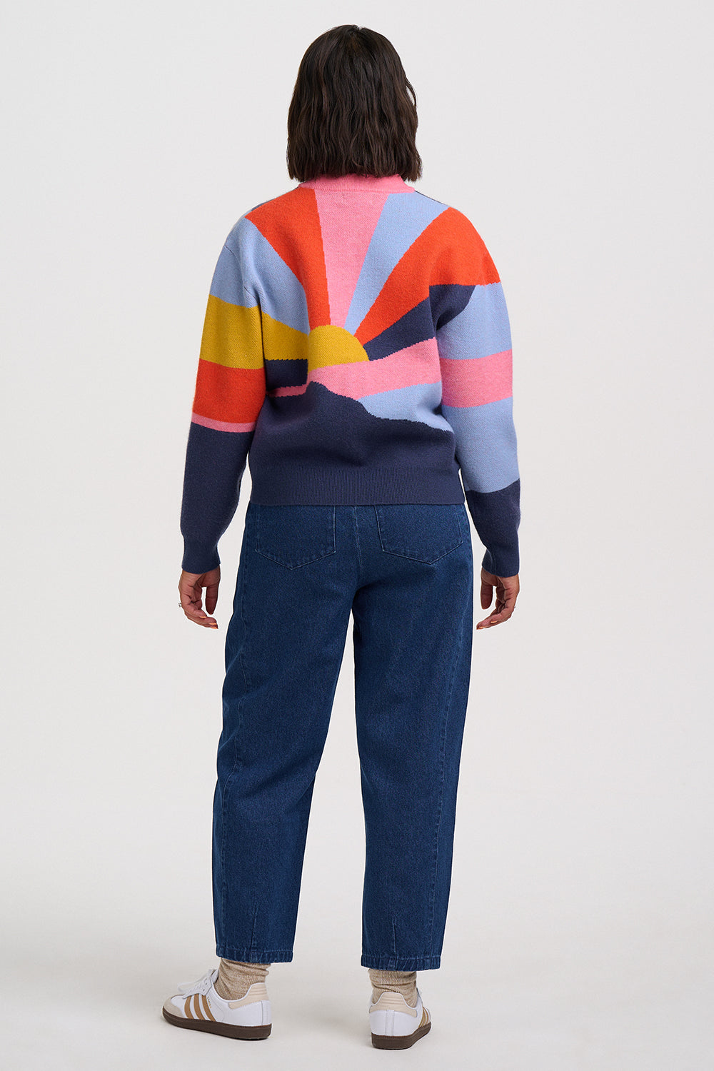 A model viewed from the back wearing a colorful striped sweater with a geometric sunburst pattern, dark blue jeans, and white sneakers, set against a plain light gray studio background.