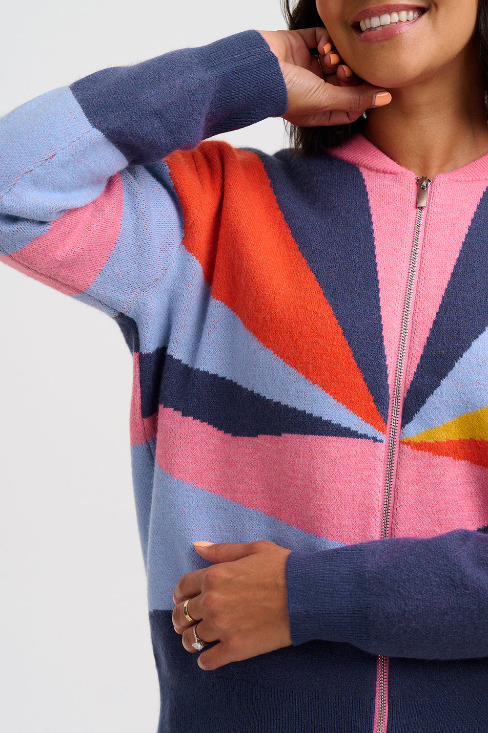 A close-up of a woman wearing a colorful zip-up sweater with geometric color-blocked panels in pink, orange, light blue, navy, and a small yellow section, against a plain light gray background.