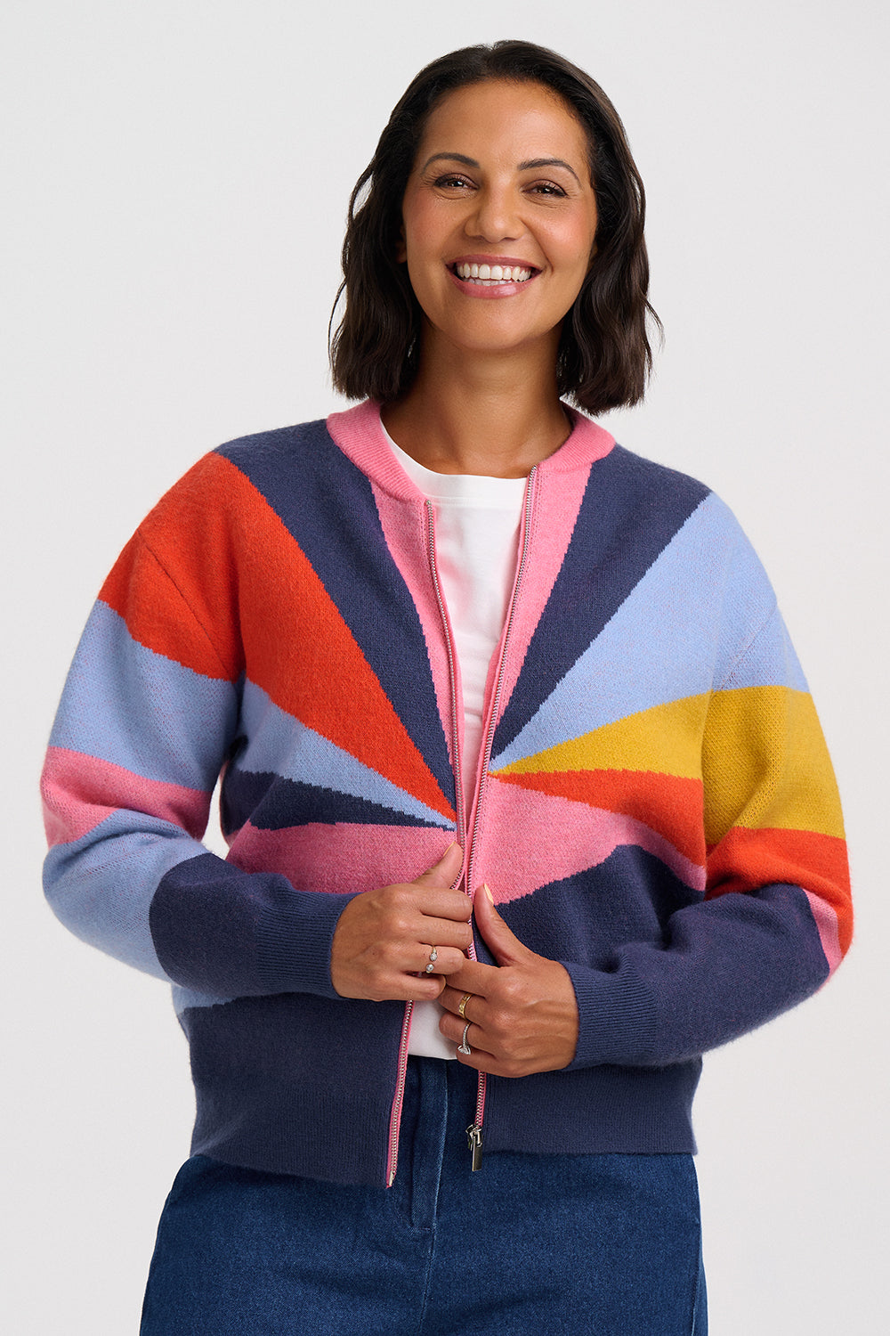 A smiling woman wearing a colorful zip-up cardigan sweater with a geometric sunburst pattern in navy, pink, orange, light blue, yellow, and purple, paired with a white top and blue jeans, against a light gray studio background.
