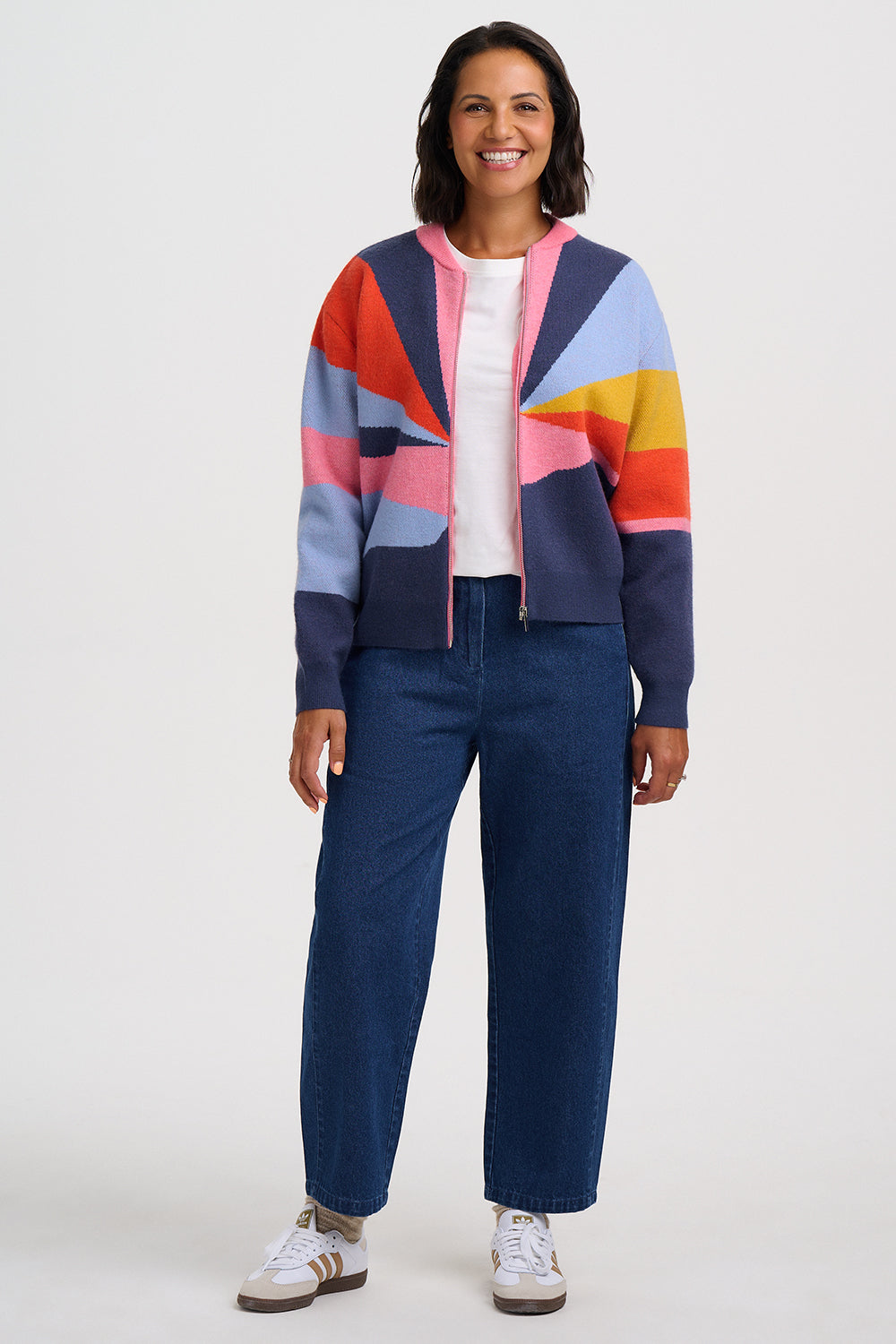 A woman modeling a colorful geometric zip-up cardigan with navy, pink, orange, yellow, and light blue panels, paired with dark blue jeans and white sneakers, against a plain light gray studio background.