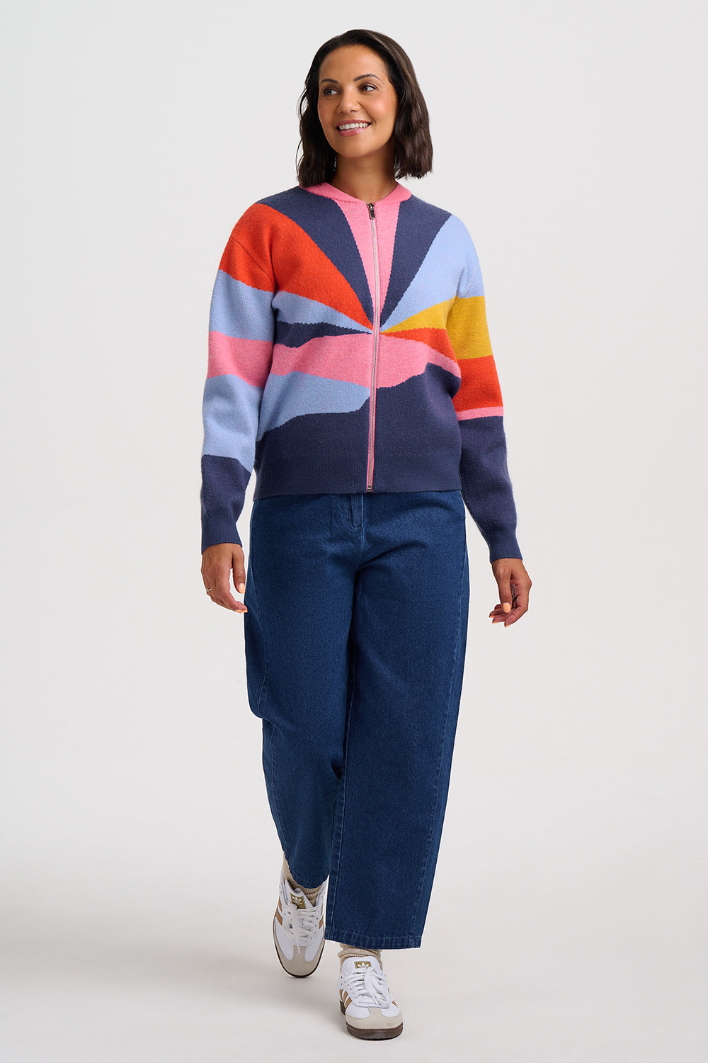 A woman modeling a colorful zip-up cardigan with a geometric stripe pattern in navy, pink, orange, light blue, and yellow, paired with dark blue jeans and white sneakers, against a light gray studio background.