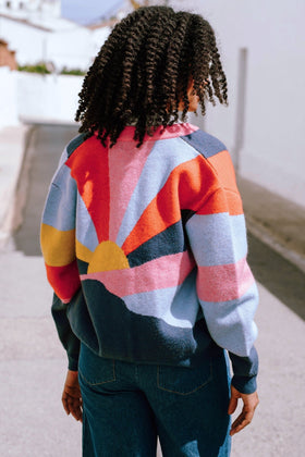A person facing away from the camera wears a multicolored knitted bomber jacket with a geometric pattern featuring shades of navy, blue, orange, pink, and mustard.