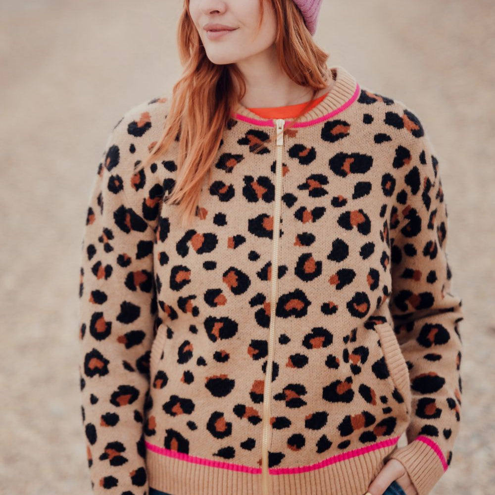 Woman wearing a leopard print jacket with pink trim and a pink beanie on a beach.