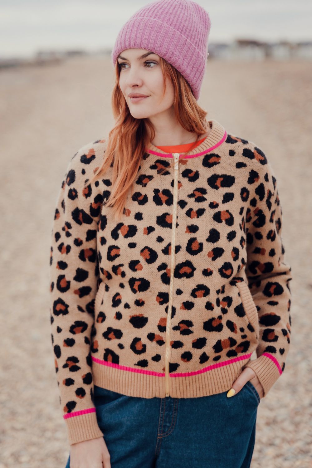 Woman wearing a leopard print jacket with pink trim and a pink beanie on a beach.