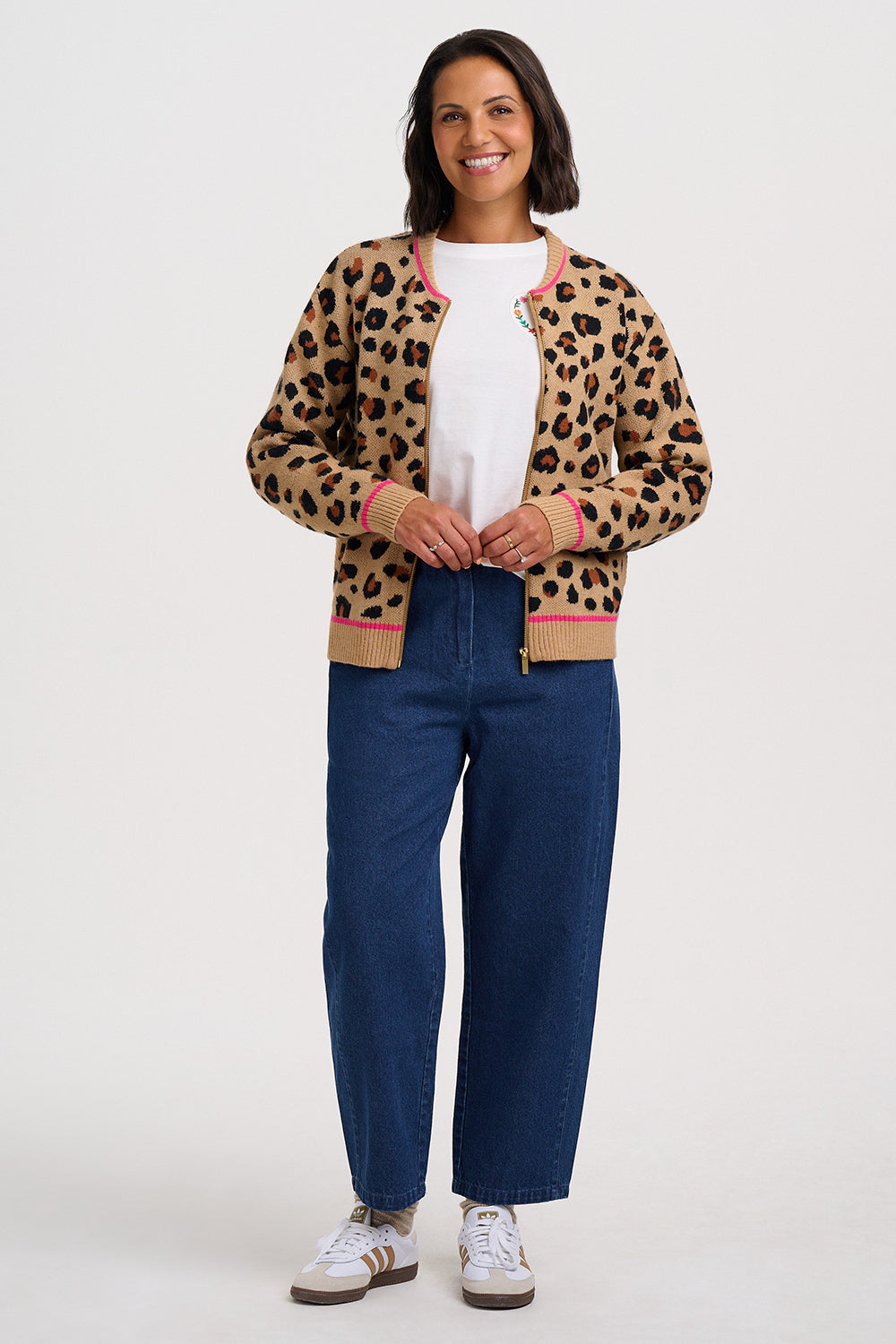 A woman modeling a tan leopard-print knit cardigan with pink and green trim, worn over a white t-shirt and dark blue jeans, standing against a light gray studio background.
