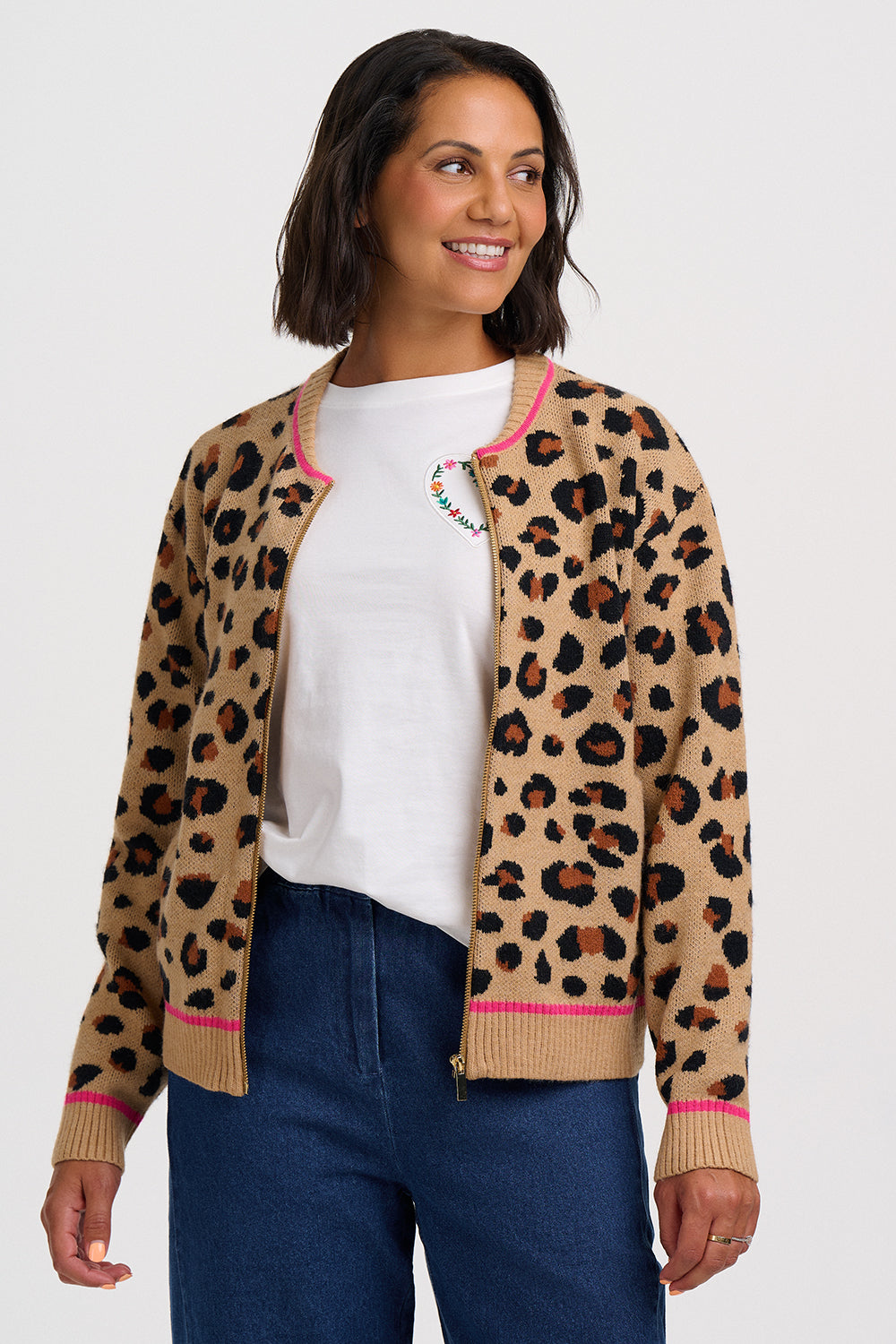 A woman wearing a beige leopard-print knit zip-up cardigan with pink trim, styled over a white t-shirt and blue jeans against a light gray studio background.
